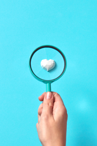 Woman's hand holds big magnifying glass above plaster figure of heart in set on a pastel blue background, copy space. Valentine's day greeting card.