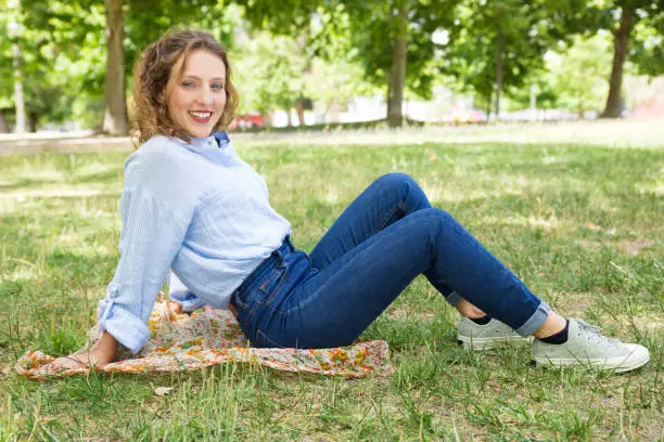 Beautiful smiling young woman sitting on headkerchief in park. Attractive lady wearing casual clothing resting in spring park. Leisure concept
