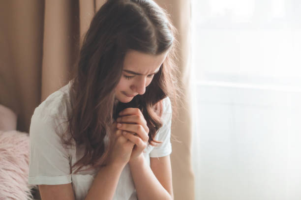 teenager girl closed her eyes, praying in a in the living room. hands folded in prayer concept for faith, spirituality and religion - praying girl imagens e fotografias de stock