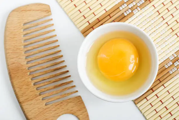 Raw egg in the small white bowl and wooden hair comb. Natural homemade hair treatment concept. Top view, copy space
