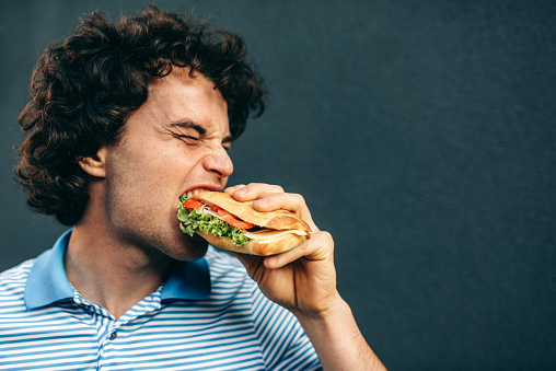Side view of close-up portrait of young handsome man eating a healthy burger. Hungry man in a fast food restaurant eating a hamburger outdoors. Man with curly hair having street food and eat a burger.