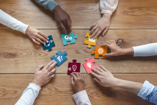 Hands of international coworkers sitting around table, putting colorful puzzles together, teamwork concept, top view