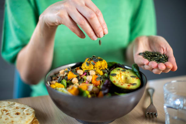 primer plano de mujer añadiendo semillas de calabaza a una comida vegana saludable en un tazón - leguminosas fotografías e imágenes de stock