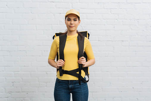 Attractive courier in yellow uniform with thermal backpack looking at camera near brick wall