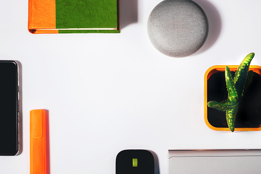 Flat lay of smart speaker with cropped smartphone, green and orange notepad, marker, aloe vera plant and laptop with a mouse
