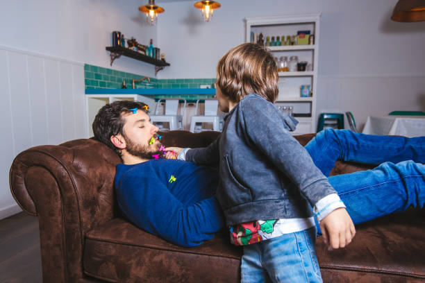 Little kid doing mischief to his father while sleeping Father's day concept. Boy placing colored clothespins on his father's beard while sleeping on the couch happy fathers day funny stock pictures, royalty-free photos & images