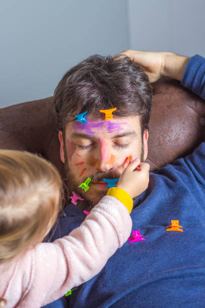 Father's Day celebration. Young man sleeping on the sofa while his little daughter makeup her face with colorful watercolors and hair clips Young father suffering the mischief of his little daughter. Little girl paints with brush and watercolors her father's face while he sleeps on the couch happy fathers day funny stock pictures, royalty-free photos & images