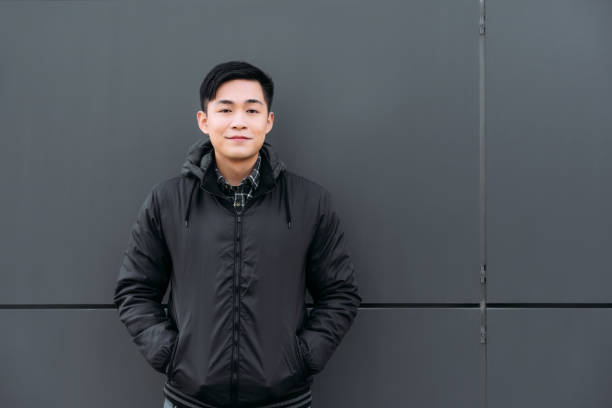 istock young asian man smiling at camera while standing by wall and holding hands in pockets 1206250642