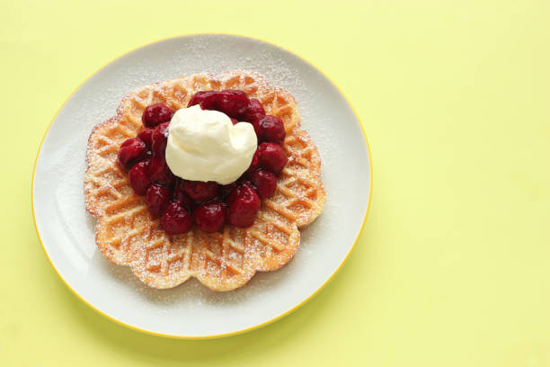 delicious waffles topped with hot cherries and cream on a white plate, yellow background with copy space