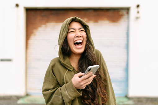 A portrait of a young and happy woman wearing a hoodie and holding a smart phone while laughing.