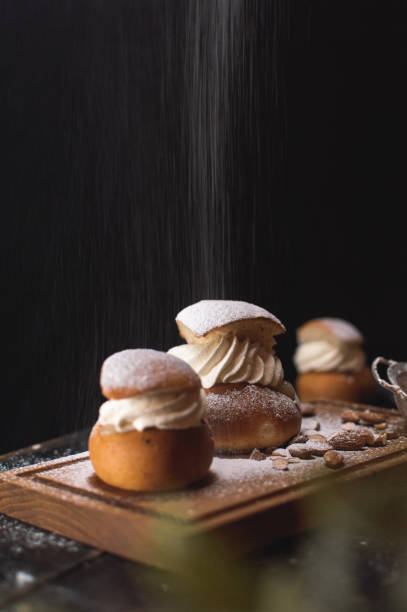 Sprinkling sugar on a Swedish semla Sprinkling sugar on a Swedish semla on a wooden cutting board next some almonds and another semla. sprinkling powdered sugar stock pictures, royalty-free photos & images