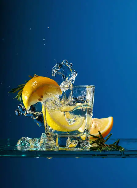Cocktail gin-tonic with lemon and rosemary. Carbonated drink with ice pieces on a glass table. Blue background, copy space.