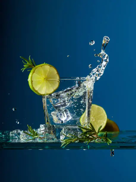 Cocktail gin-tonic with lime and rosemary. Carbonated drink with ice pieces on a glass table. Blue background, copy space.