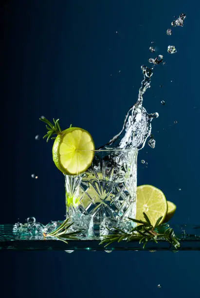 Cocktail gin-tonic with lime and rosemary. Carbonated drink with ice pieces on a glass table. Blue background, copy space.