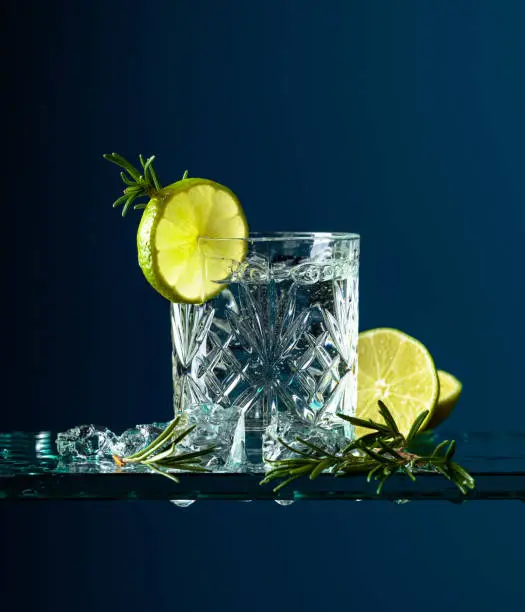 Cocktail gin-tonic with lime and rosemary. Carbonated drink with ice pieces on a glass table.