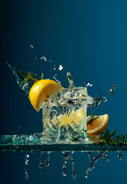 Cocktail gin-tonic with lemon and rosemary. Carbonated drink with ice pieces on a glass table.