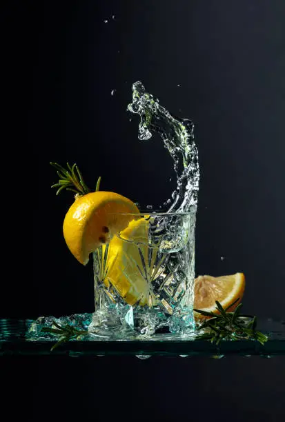 Cocktail gin-tonic with lemon and rosemary. Carbonated drink with ice pieces on a glass table. Black background, copy space.
