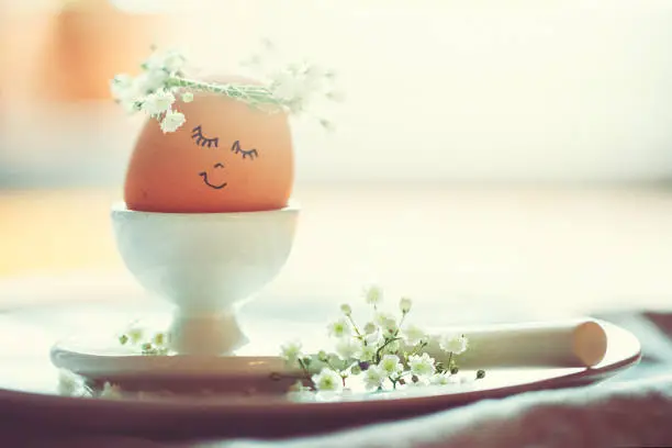 Natural easter egg in an eggcup with funny painted face and sweet flower wreath on Easter breakfast