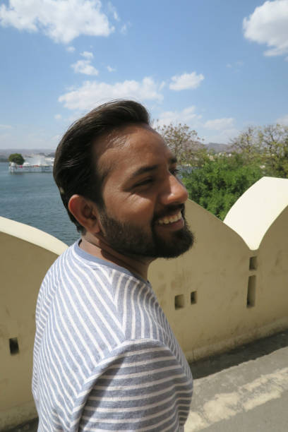 Stock photo of young happy Hindi Indian man in early 20s looking sideways, laughing with short brown beard and hair brushed back quiff, close-up portrait of good-looking guy from Delhi, India, wearing blue and white striped T-shirt walking outdoors on bridge imagen del joven joven indio hindi feliz a principios de los años 20 riendo mirando a la distancia, barba marrón corta y el pelo cepillado de plumas hacia atrás, retrato de cerca de un tipo guapo de delhi, india, vestido azul y blanco rayas camiseta cam