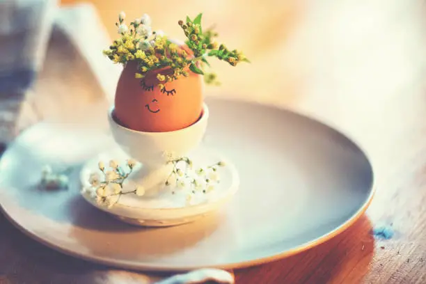 Natural easter egg in an eggcup with funny painted face and sweet flower wreath on Easter breakfast