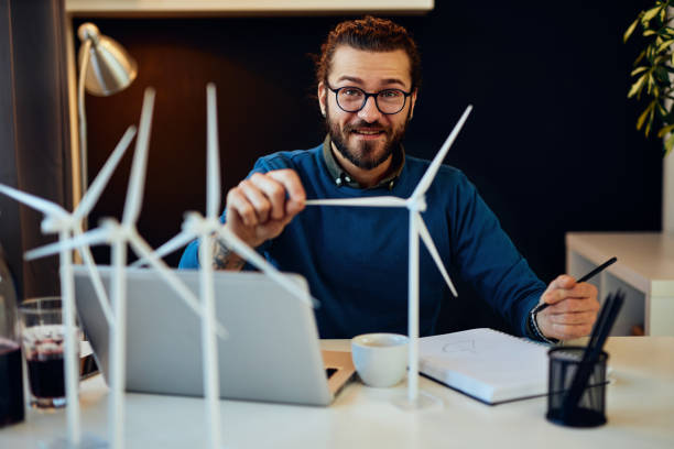 istock Young creative caucasian bearded environmentalist  with curly hair sitting in his office, touching windmill model and drawing new better model in notebook. 1206272821