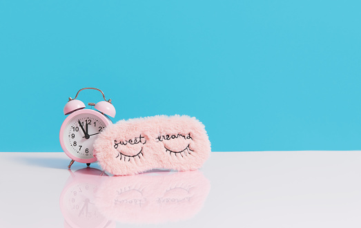 Sleep optimization. Pink fluffy mask for sleeping and a pink alarm clock on a light blue background.