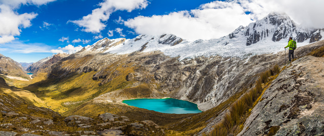Male Adventurer with a Backpack on top of a hill on the right looking at the beautiful and vast valley landscape with a turquoise blue water color lake in Cordillera Blanca, Peruvian Mountains, South America