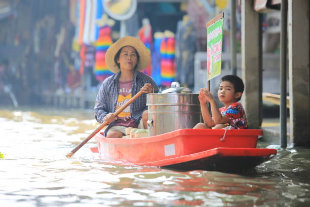 Group of tourists in tourboat in Amphawa captured on almost water level of river.  Boat is anchored in old part of Amphawa with floating market area and many shops and restaurants in old wooden houses. Tourboats are making trips along Mae Khlong river around Amphawa. People are wearing life jackets