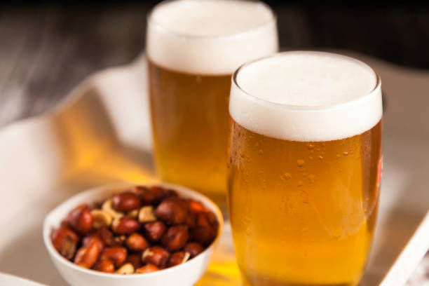 due bicchieri di birra con una ciotola di arachidi in un vassoio - beer nuts foto e immagini stock