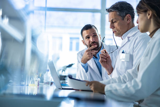 Team of scientists cooperating while working on laptop in laboratory. Group of biotechnologists cooperating while reading scientific data on a computer in laboratory. Focus on thoughtful man. researcher and doctor stock pictures, royalty-free photos & images