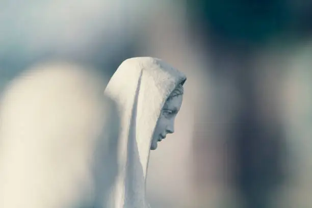 White cemetery representation of Mary, mother of Jesus, in a Surrey cemetery in winter sunlight, picked out and isolated through selective focus with a long focus / 400mm lens. Copy space.