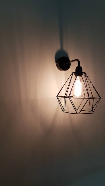 istock Minimal style pendant light closeup. Diamond shape wire lampshade with light bulb inside. White glowing filament. Sconce lamp hanging on wall background with shadows and copy space. Interior lighting 1206403011