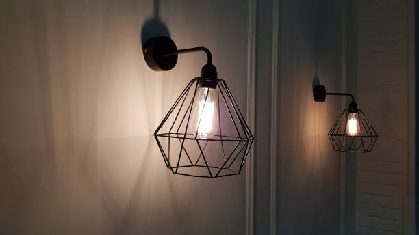 istock Geometric pendant light closeup on blurry background. Diamond shape wire lampshade with light bulb inside. Sconce lamps hanging on wall background with shadows and copy space. Interior lighting. 1206403039