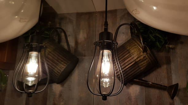 istock Industrial style wire lampshades with shining light bulb inside and grunge background of blurry silhouettes of watering cans. Glowing filament inside lamps hanging in front of shabby wall. 1206403082