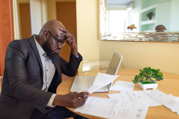 istock Shot of afro american young man in a home office using laptop and taking notes. Black guy sitting at table and working from home office. Worried Mature African is Looking at Financial Bills at Home Office 1206405875