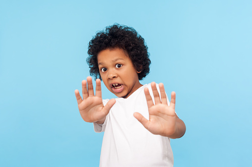 Child phobia. Portrait of scared little boy looking terrified panicked at camera and showing stop gesture as if trying to defend himself, screaming in fear. studio shot isolated on blue background