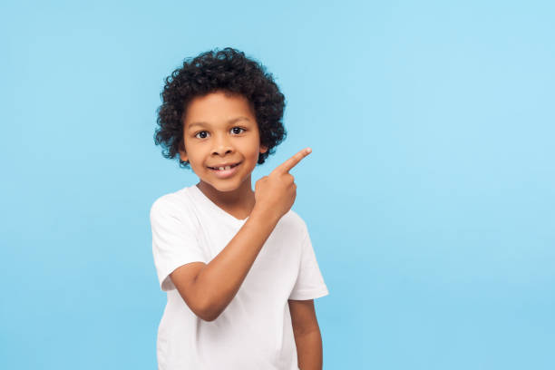 見て、ここで広告!白いtシャツで巻き毛のあるかわいい陽気な小さな男の子の肖像画が空の場所を指し示す - child ideas inspiration expertise ストックフォトと画像