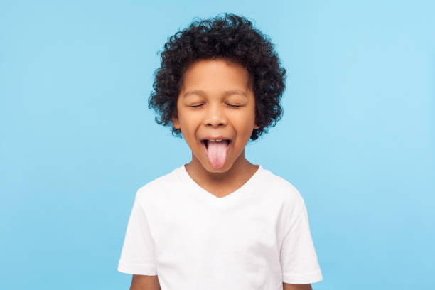 porträt von lustigen freche kleine junge mit lockigen haaren in t-shirt herausragend zunge und halten die augen geschlossen, ungehorsamkind - coole jungs frisuren stock-fotos und bilder