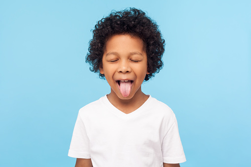 Portrait of funny naughty little boy with curly hair in T-shirt sticking out tongue and keeping eyes closed, disobedient child foolishing with cute derisive expression. studio shot blue background