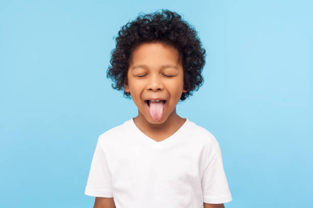 Portrait of funny naughty little boy with curly hair in T-shirt sticking out tongue and keeping eyes closed, disobedient child Portrait of funny naughty little boy with curly hair in T-shirt sticking out tongue and keeping eyes closed, disobedient child foolishing with cute derisive expression. studio shot blue background making a face stock pictures, royalty-free photos & images