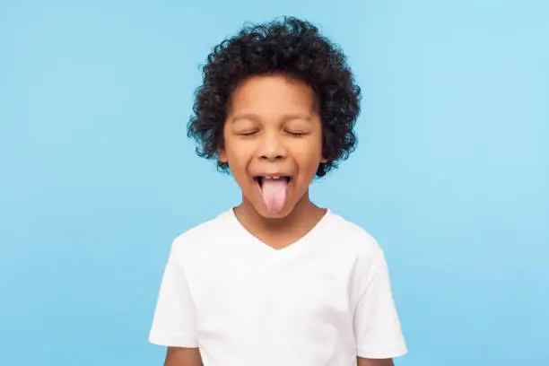 Portrait of funny naughty little boy with curly hair in T-shirt sticking out tongue and keeping eyes closed, disobedient child foolishing with cute derisive expression. studio shot blue background