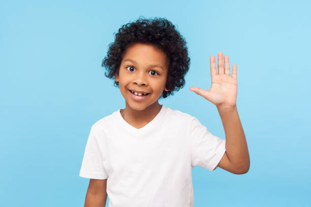 Portrait of amazing friendly little boy grinning happily and waving hi, gesturing hello with raised arm, welcoming greeting Portrait of amazing friendly little boy grinning happily and waving hi, gesturing hello with raised arm, welcoming greeting, expressing joy from meeting. indoor studio shot isolated on blue background kid waves stock pictures, royalty-free photos & images