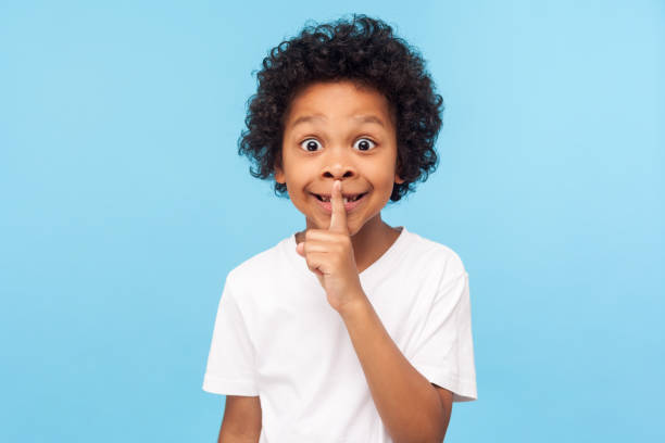shh, be quiet! portrait of funny cute little boy with curly hair in t-shirt making silence gesture with finger on his lips - segredo criança imagens e fotografias de stock
