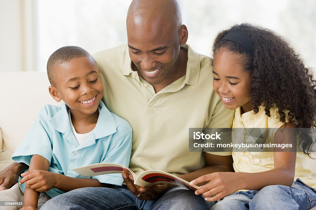 Man sits and reads a book with two children Man and two children sitting in living room reading book together and smiling Reading Stock Photo