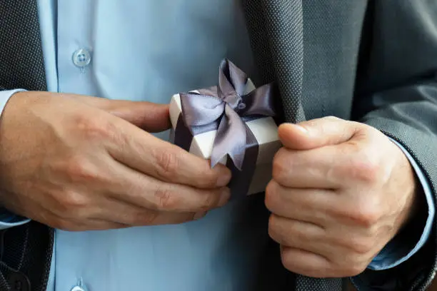 Chest view of an unrecognizable businessman wearing a suit who is about to give a gift box out of his jacket. The white gift box is wrapped with purple ribbon.