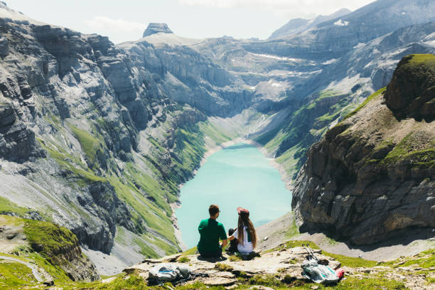 Young woman and man backpackers getting up to the top of the mountains with scenic view of turquoise colored lake in Swiss Alps, embracing loving each other and looking at the beautiful panoramic view in Switzerland