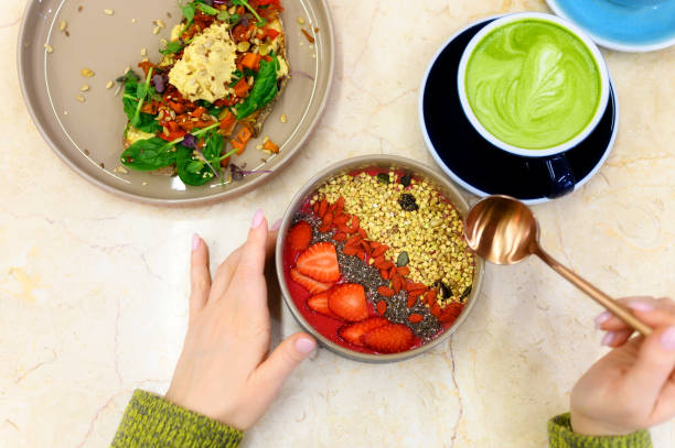 smoothie bowl with strawberry, goji, and green buckwheat granola, and matcha latte cup, and toast with hummus, and female hands with golden spoon, ready to eats. raw healthy food concept - food vegan food gourmet vegetarian food imagens e fotografias de stock