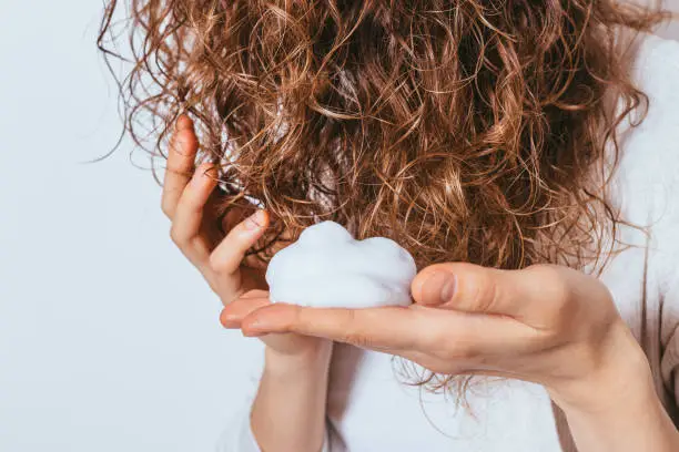 Woman's hands apply styling mousse to her curly hair on white studio background, close-up.