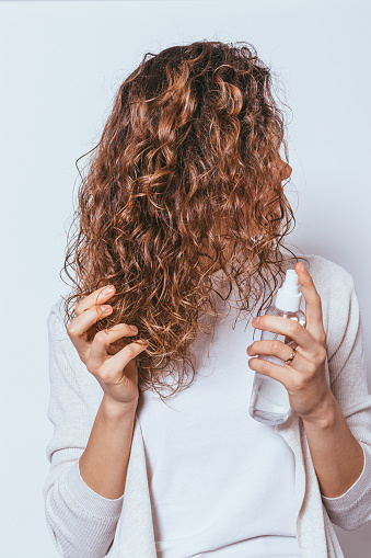 Young woman applying moisturizing spray to her curly hair on white studio background.