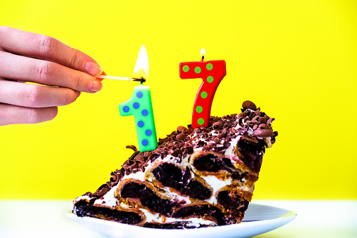 Birthday cake, 17 years old. A piece of cake. In the hand a match lighting candles. Yellow background.
